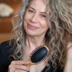 Smiling woman with natural grey curly hair holding a hairbrush, showcasing a stylish and elegant curly hairstyles for women over 50.